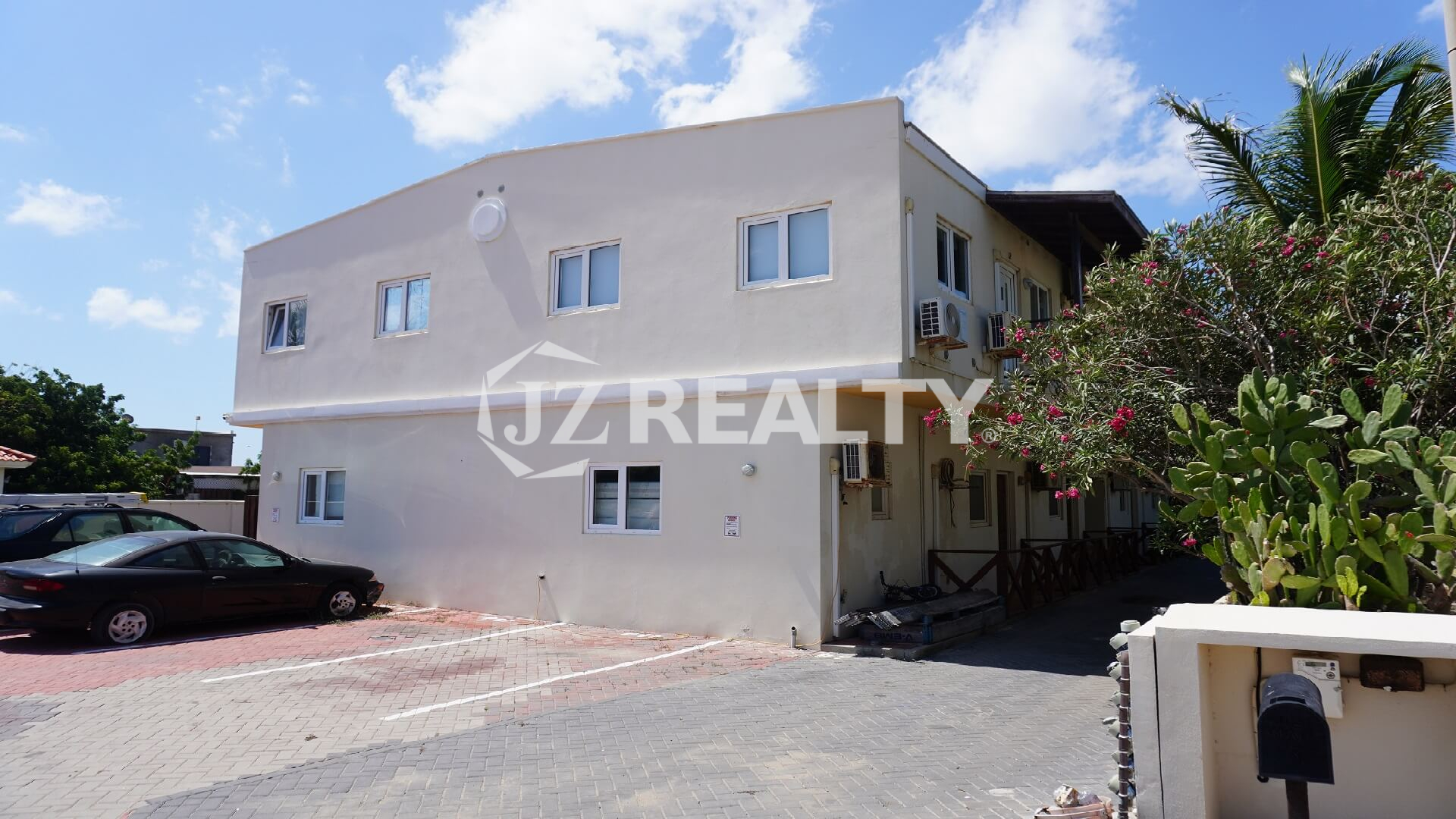 Exterior of two-story beige apartment building with private parking and garden plants.