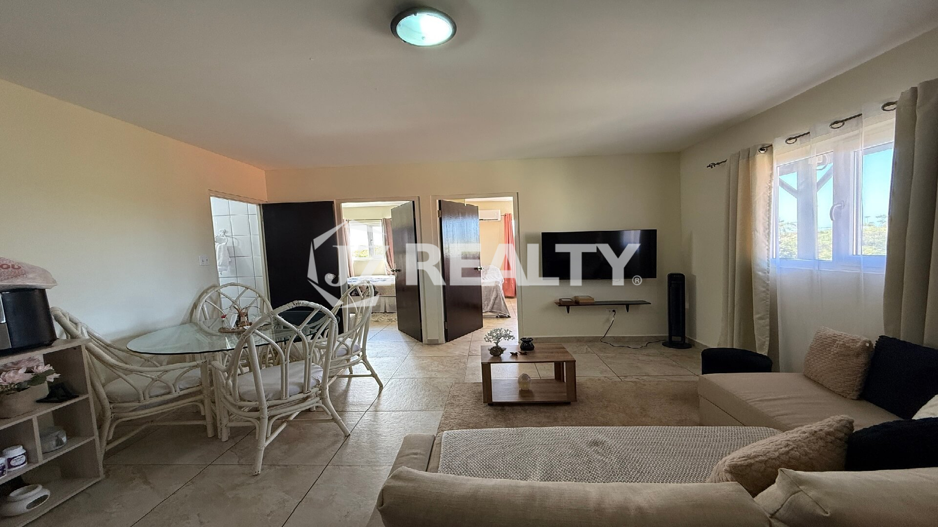 Living area showing a mounted television, large window with curtains, and doors leading to bedrooms.