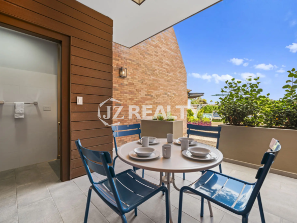 Porta al Sole Unit 11 outdoor patio with a round white dining table, four blue chairs, and a brick wall backdrop under a blue sky.