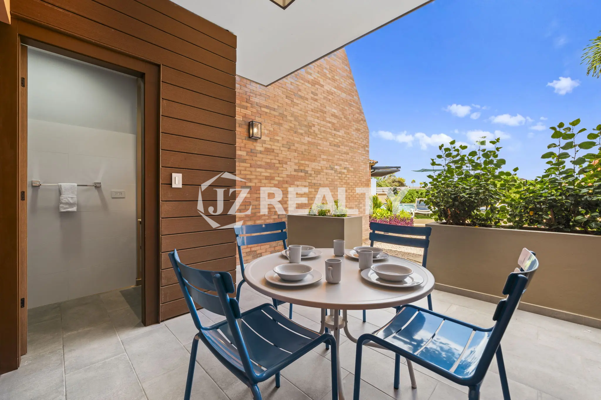 Porta al Sole Unit 11 outdoor patio with a round white dining table, four blue chairs, and a brick wall backdrop under a blue sky.