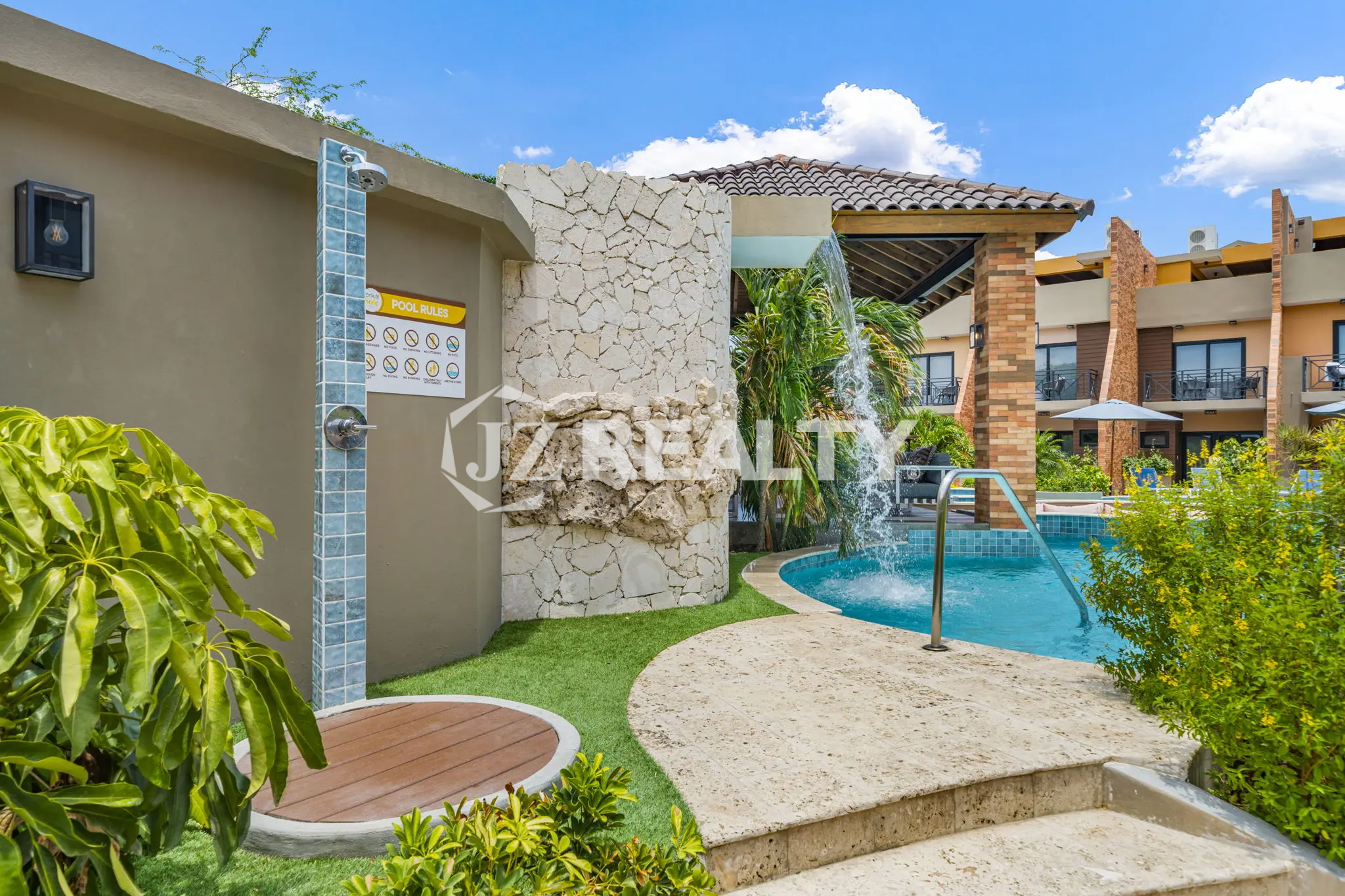 Porta al Sole Unit 11 outdoor shower station next to the pool, featuring a stone rock wall, wood-slat circular base, and pool rules sign.