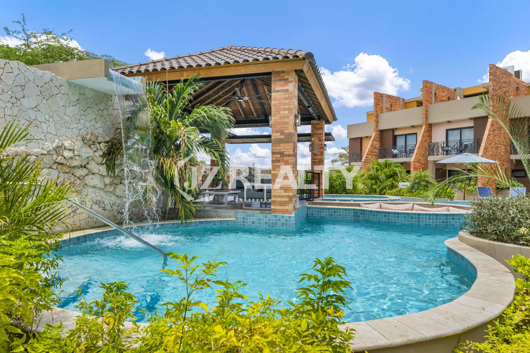 Porta al Sole Unit 11 community pool area with a decorative rock waterfall, covered lounge gazebo, and integrated shallow spa/jacuzzi area.
