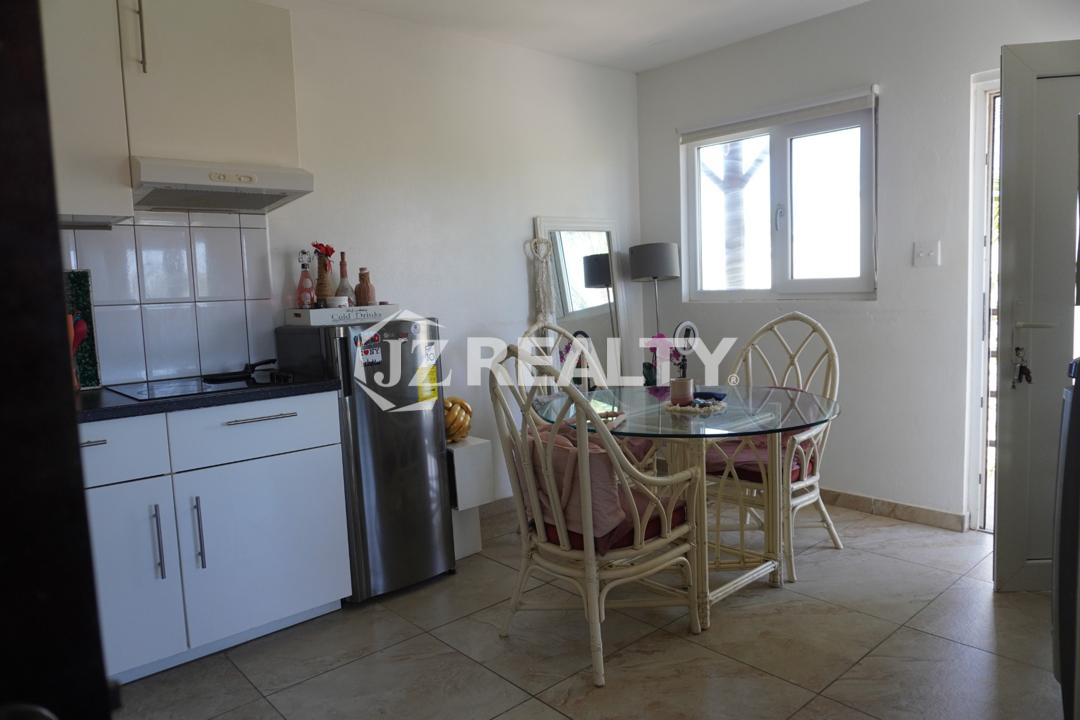 Dining area with glass table and rattan chairs next to kitchen and window in an Aruba apartment.