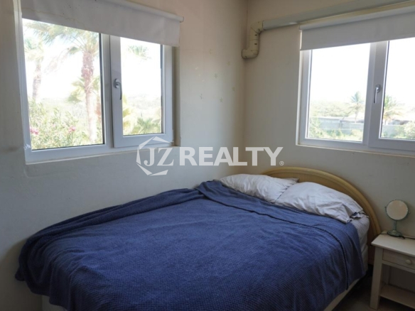 Bright bedroom with corner windows, blue bedspread, and nightstand in an Aruba apartment.