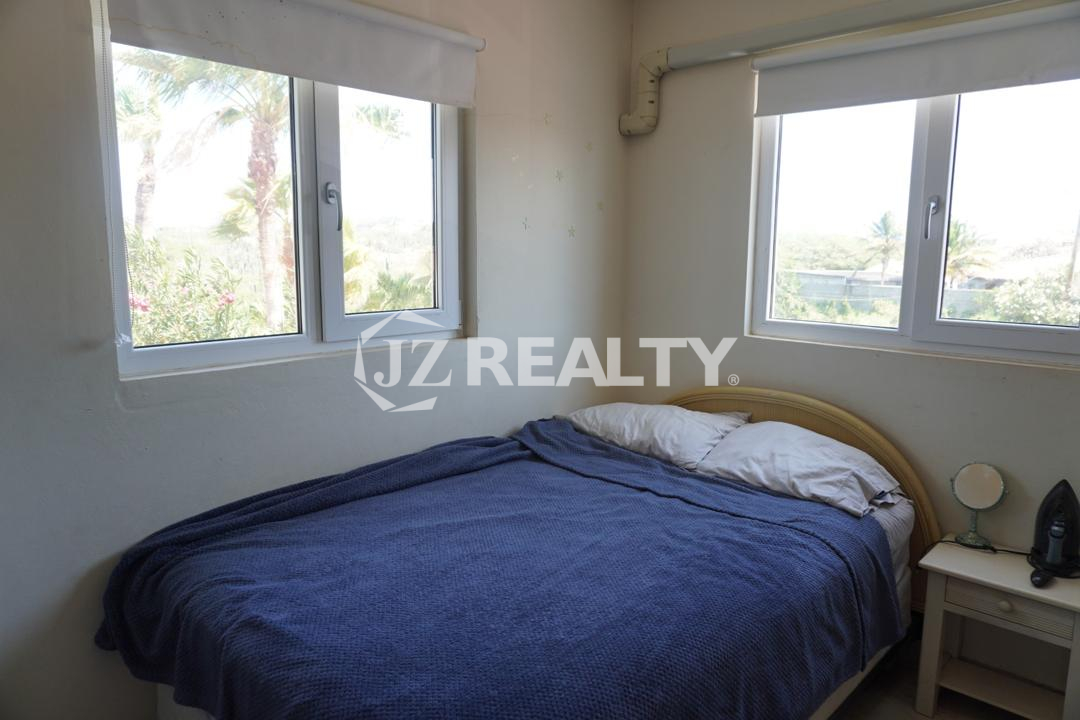 Bright bedroom with corner windows, blue bedspread, and nightstand in an Aruba apartment.
