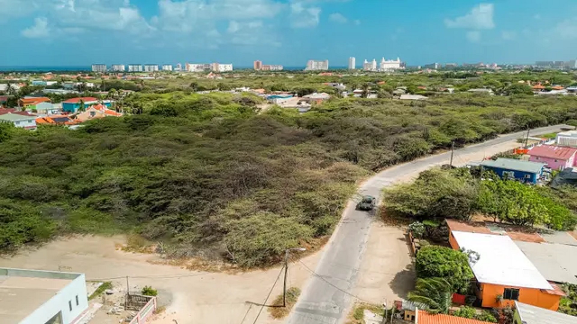 Wide aerial overview of development land for sale in Bubali, Noord, Aruba, featuring nearby homes, vegetation, and distant urban areas.