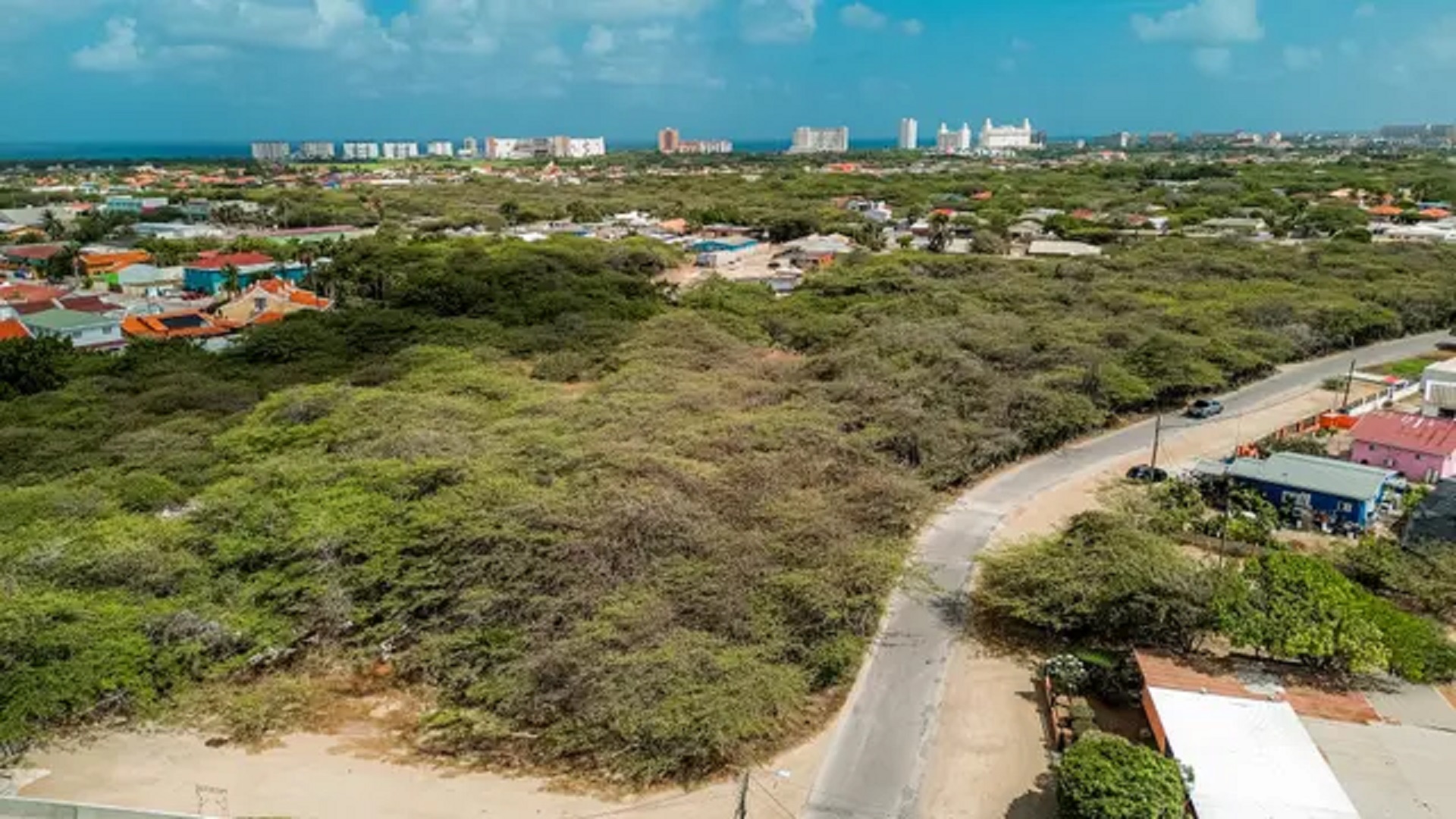 Aerial view of Bubali development land in Noord, Aruba, featuring a curved road, surrounding vegetation, and distant skyline.