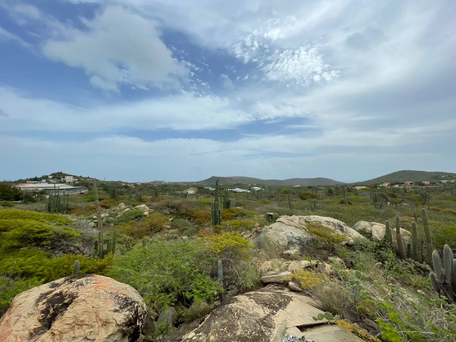 Ground-level view of the land for sale in Mon Pos, Santa Cruz, Aruba, showing natural terrain with rock formations, cacti, and surrounding hills.