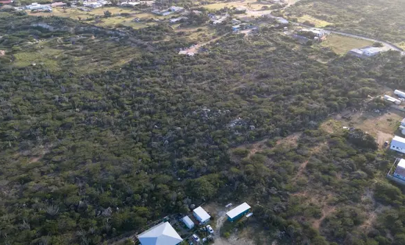 Broad aerial view of the land for sale in Mon Pos, Santa Cruz, Aruba, with neighboring structures and open terrain nearby.