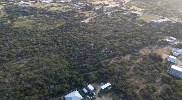 Broad aerial view of the land for sale in Mon Pos, Santa Cruz, Aruba, with neighboring structures and open terrain nearby.
